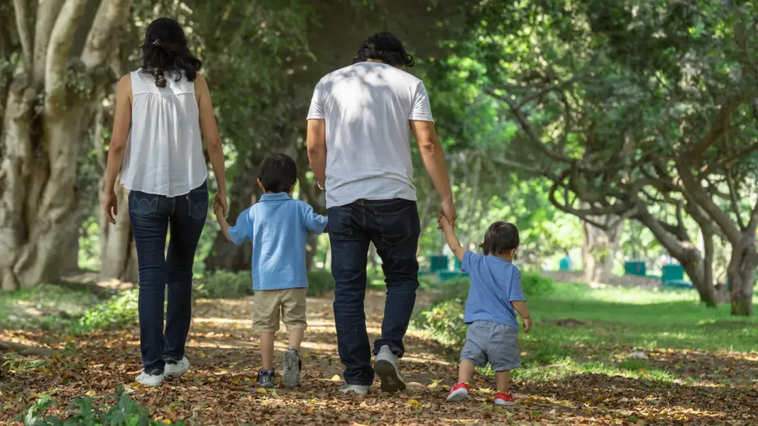 Família com dois adultos e duas crianças caminhando de mãos dadas por um parque arborizado