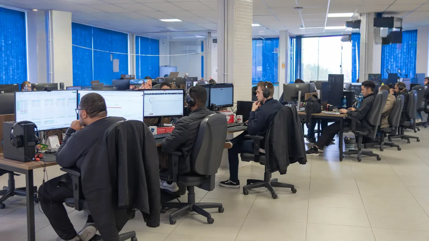 Sala de trabalho da Conplan: fileiras de estações com dois monitores cada e a equipe atendendo de fone de ouvido, junto às janelas