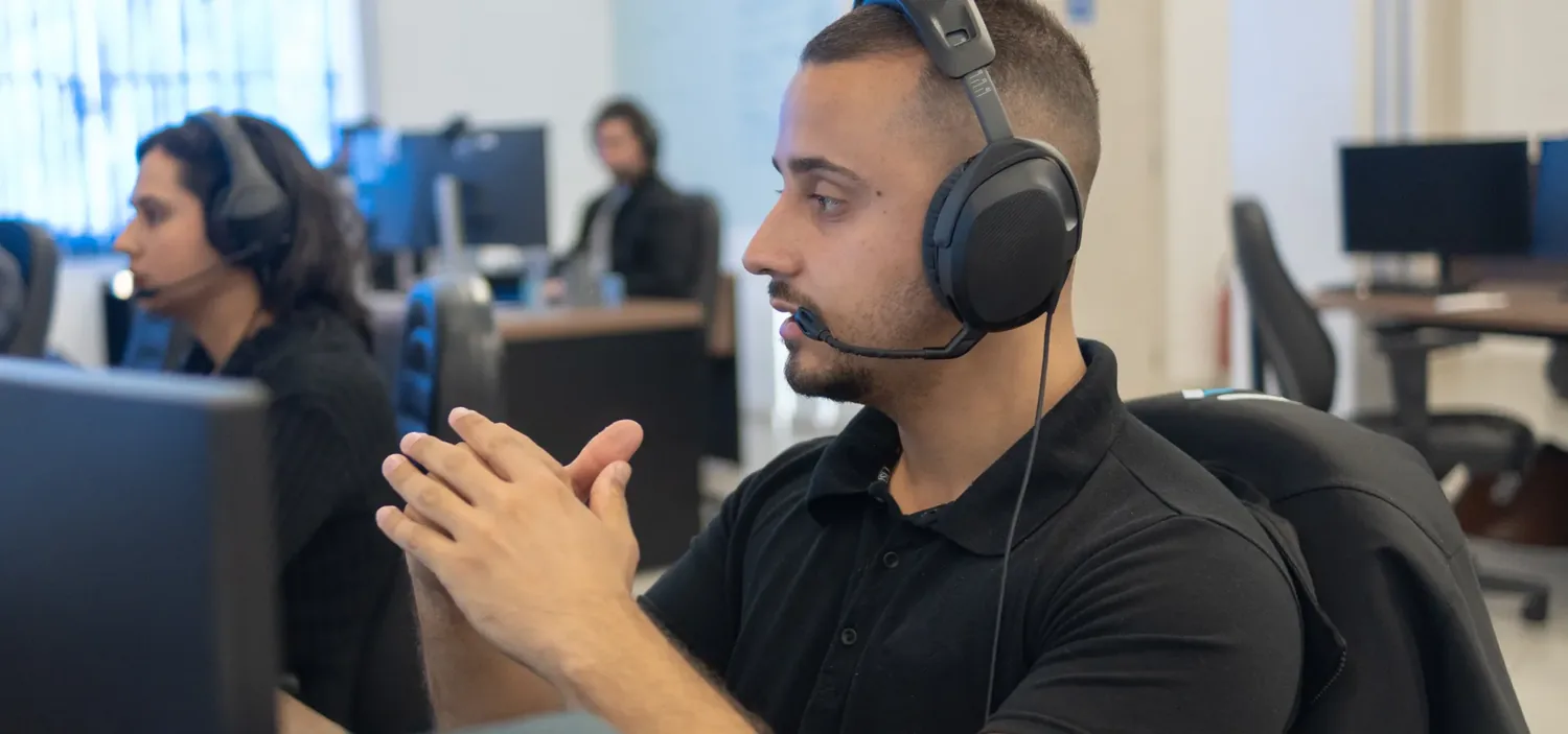 Analista da Conplan com fone de ouvido conversando na esta��o de trabalho, com a equipe ao fundo